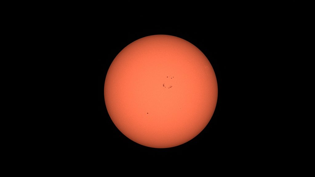Photo of the sun through a solar filter, a black background with an orange sun showing sunspots near the centre of the disk
